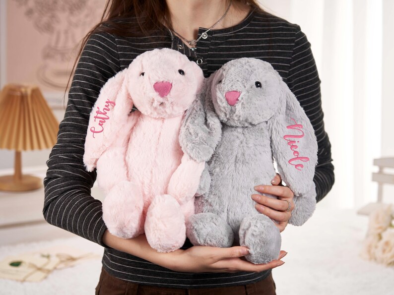 May include: Two plush bunny rabbits, one pink and one gray, are held in front of a person. The pink bunny has the name "Cathy" embroidered on its ear, and the gray bunny has "Nicole" embroidered on its ear. Both bunnies have pink noses.