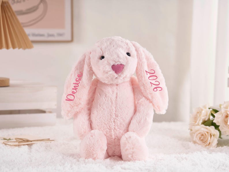 May include: A soft, pink plush bunny with long ears and a heart-shaped nose. The name "Denise" and the year "2026" are embroidered on the ears. The bunny is sitting on a white surface.