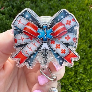 May include: A badge reel with a bow-shaped design featuring medical symbols. The bow has red, white, and blue ribbons with a heartbeat pattern and red crosses. A blue Star of Life symbol is at the center, with a caduceus below.
