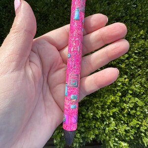 May include: A pink glitter pen with a black tip and silver accents. The pen is decorated with small, colorful illustrations of medical tools and equipment. The pen is held in a hand.