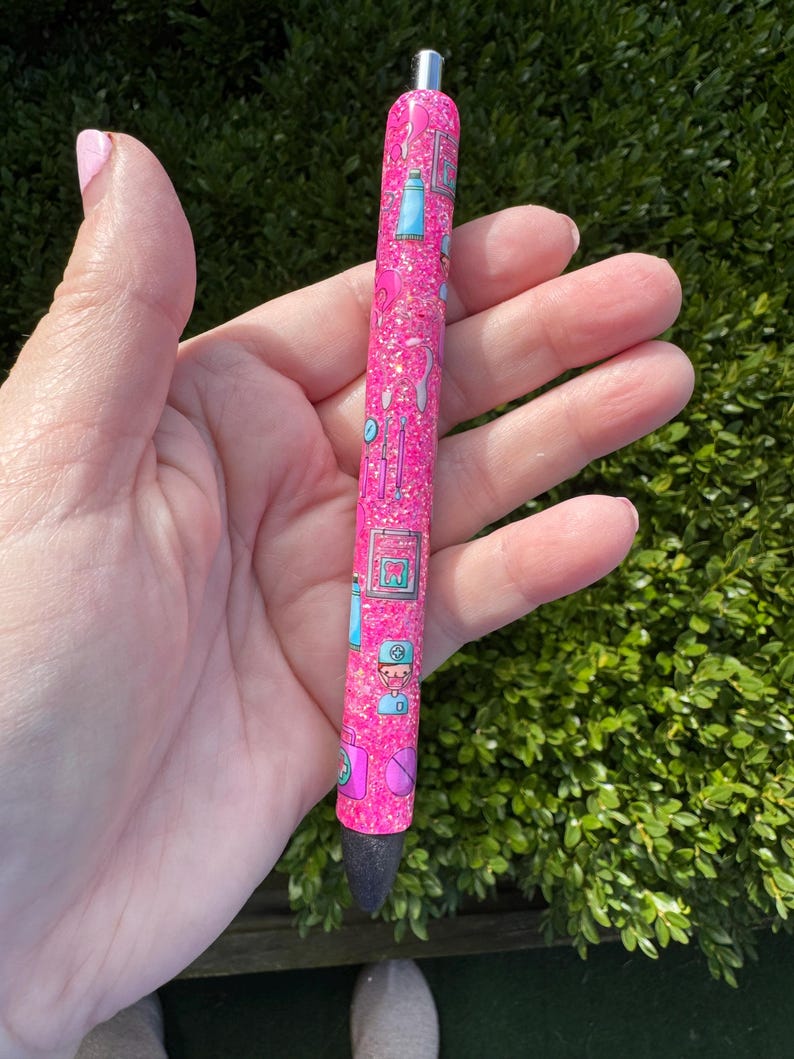 May include: A pink glitter pen with a black tip and silver accents. The pen is decorated with small, colorful illustrations of medical tools and equipment. The pen is held in a hand.