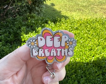 Op de afbeelding: Een badge reel met de woorden "DEEP BREATHS" in een bubble-stijl lettertype. Het ontwerp heeft een roze achtergrond met een gele en oranje omtrek, en kleine witte bloemen. De reel heeft een doorzichtige plastic band en een metalen clip.