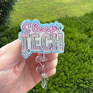 May include: A badge reel with the words "Sleep TECH" in pink and blue glitter lettering. The badge has a light blue glitter border and a clear plastic clip. The background is a green, leafy area.