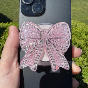 May include: A black smartphone with a pink glitter bow phone grip attached. The bow has a black outline and is held in a person's hand. The background is green grass.
