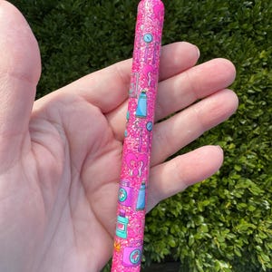 May include: A pink glitter pen with a pattern of medical-themed illustrations, including hearts, first aid kits, and medicine bottles. The pen has a silver tip and a black writing end. The pen is held in a hand against a green leafy background.