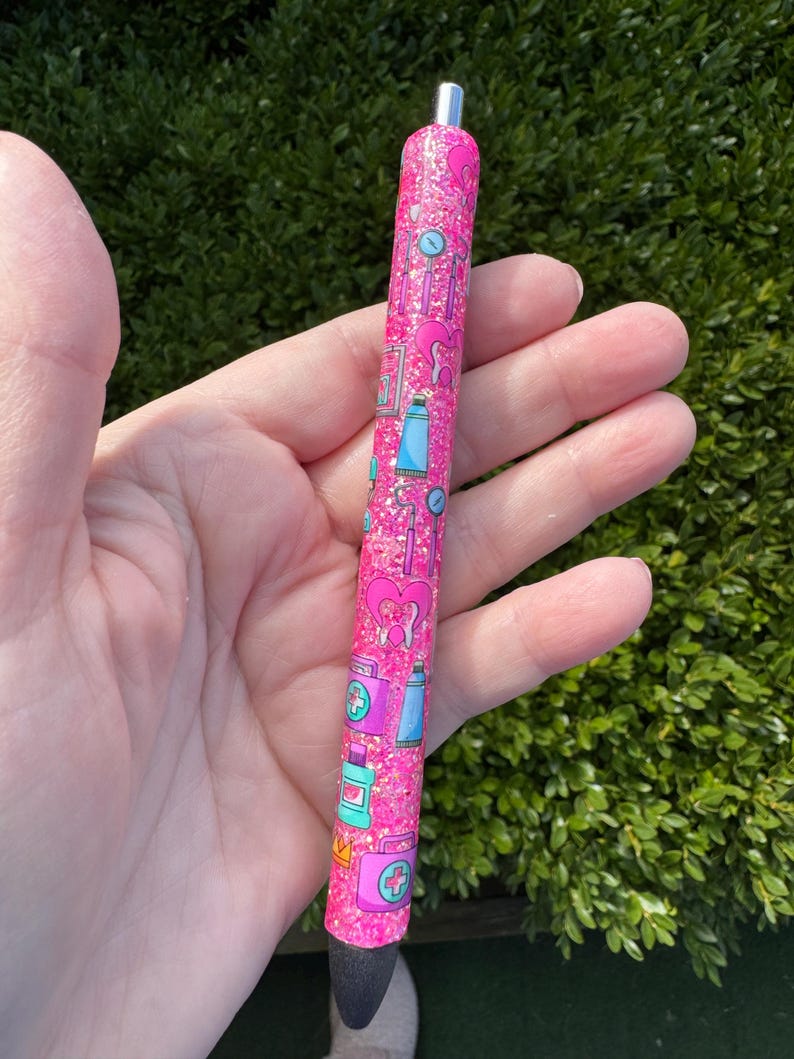 May include: A pink glitter pen with a pattern of medical-themed illustrations, including hearts, first aid kits, and medicine bottles. The pen has a silver tip and a black writing end. The pen is held in a hand against a green leafy background.