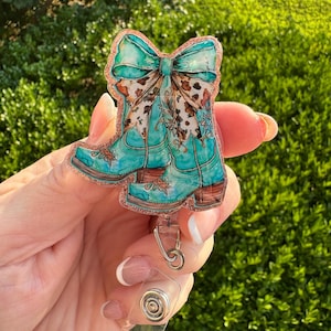 May include: A turquoise badge reel featuring a cowboy boot design with a bow. The bow has a brown and white cow print pattern. The badge reel has a clear plastic clip and a retractable cord.