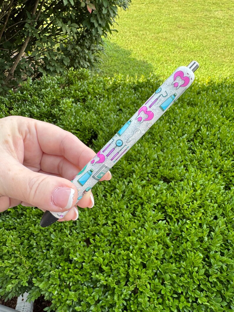 May include: A pen with a white glittery barrel featuring pink and blue dental-themed graphics. The pen has a black tip and a silver metal top. The design includes tooth, toothbrush, and dental tool illustrations.