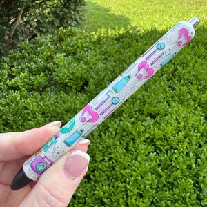May include: A glittery pen with a black tip and silver accents. The pen is decorated with dental-themed illustrations in pink, teal, and purple. The pen is held against a backdrop of green foliage.