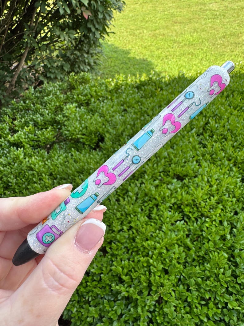 May include: A glittery pen with a black tip and silver accents. The pen is decorated with dental-themed illustrations in pink, teal, and purple. The pen is held against a backdrop of green foliage.