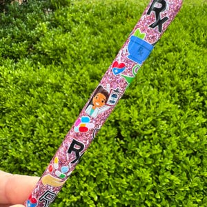 May include: A pink glitter pen with medical-themed stickers, including the Rx symbol, pills, and a doctor. The pen has a silver tip and is held against a backdrop of green foliage.