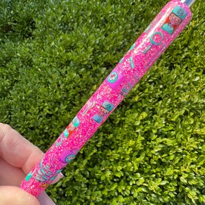 May include: A pink glitter pen with a silver tip, decorated with medical-themed illustrations. The pen features images of nurses, medical symbols, and tools. The pen is held against a backdrop of green foliage.