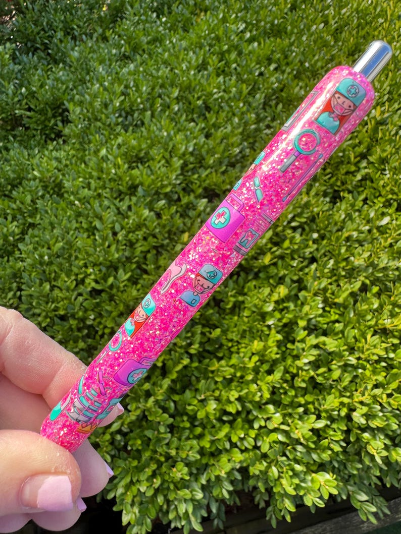 May include: A pink glitter pen with a silver tip, decorated with medical-themed illustrations. The pen features images of nurses, medical symbols, and tools. The pen is held against a backdrop of green foliage.