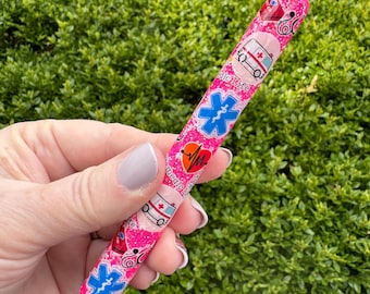 May include: A pink pen with a medical theme, featuring illustrations of ambulances, stethoscopes, and the Star of Life symbol. The pen has a glittery finish and a silver tip. The words "medic life" are also visible on the pen.