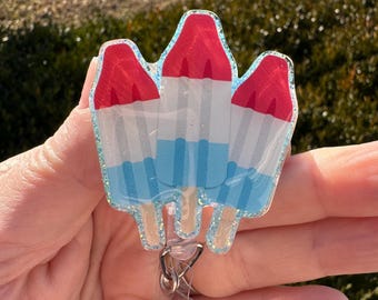 May include: A badge reel featuring a design of three red, white, and blue popsicle treats. The badge reel has a clear plastic retractable cord and a metal clip for attaching to clothing or accessories. The popsicles have a glittery outline.