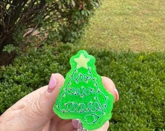 May include: A green Christmas tree badge reel with a star on top and a stethoscope design. The badge reel is held in a hand against a background of green foliage.