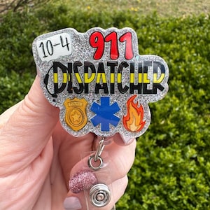 May include: A glittery badge reel with the words "911 DISPATCHER" in black and yellow, with the numbers "10-4" and "911" in red. The badge also features a gold star, a blue star of life, and a flame.