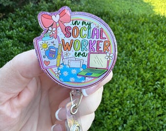 May include: A badge reel with a glittery pink border and a design that reads "in my SOCIAL WORKER era." The design includes a pink bow, flowers, a laptop, and other social work-related icons. The reel has a clear plastic clip.