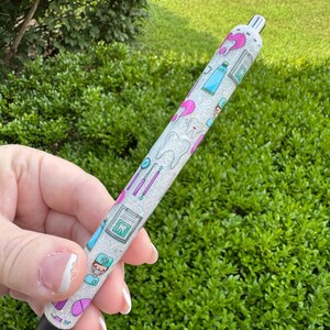 May include: A glittery white pen featuring dental-themed illustrations in pink, blue, and green. The design includes images of teeth, toothbrushes, and dental tools. The pen has a black tip and a silver metal top.