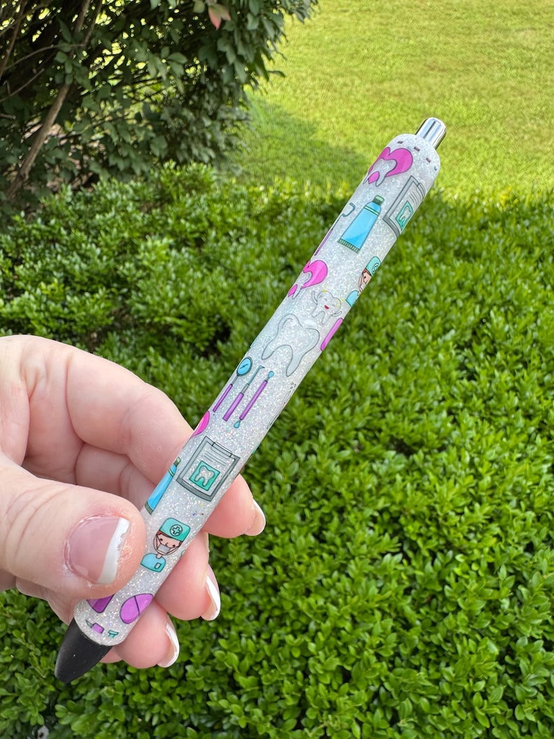 May include: A glittery white pen featuring dental-themed illustrations in pink, blue, and green. The design includes images of teeth, toothbrushes, and dental tools. The pen has a black tip and a silver metal top.