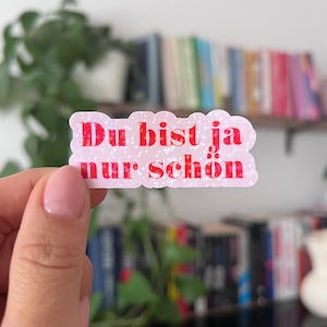 Könnte beinhalten: Ein rosa-weißer Aufkleber mit dem deutschen Text "Du bist ja nur schön" in Rot. Der Aufkleber hat ein glitzerndes, strukturiertes Aussehen und wird in einer Hand gehalten. Der Hintergrund umfasst ein Bücherregal und Grün.