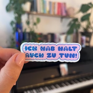 Könnte beinhalten: Ein Aufkleber mit dem Text "ICH HAB HALT AUCH ZU TUN!" in blauen Buchstaben auf rosa Hintergrund. Der Aufkleber hat einen weißen Rand mit Glitzerakzenten. Der Aufkleber wird vor einem Klavier gehalten.