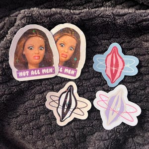 Könnte beinhalten: Eine Sammlung von Aufklebern mit Puppengesichtern und dem Text "NOT ALL MEN". Die anderen Aufkleber zeigen stilisierte Vulva-Designs in verschiedenen Farben mit holografischem Finish.