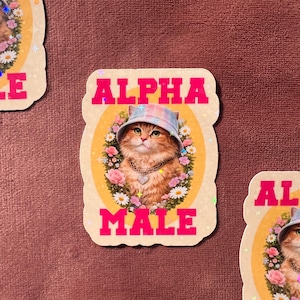 Könnte beinhalten: Aufkleber mit einer Cartoon-Katze, die einen Fischerhut und eine Halskette trägt, umgeben von Blumen. Die Worte "ALPHA MALE" stehen in rosa Blockbuchstaben über und unter dem Katzenbild. Der Aufkleber hat einen hellbraunen Rand.