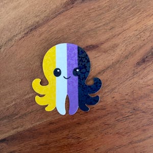Könnte beinhalten: Ein farbenfroher Oktopus-Aufkleber mit dem Design der nicht-binären Pride-Flagge. Der Oktopus hat einen gelben Kopf, einen weißen Streifen, einen lila Streifen und einen schwarzen, glitzernden Abschnitt. Der Oktopus hat ein lächelndes Gesicht und liegt auf einer Holzoberfläche.