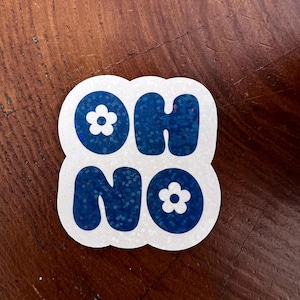 Puede incluir: Una pegatina blanca con las palabras "OH NO" en letras azules llenas de purpurina. Pequeños gráficos de flores blancas se colocan encima y debajo de las letras. La pegatina está sobre una superficie de madera.