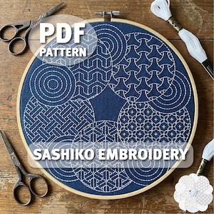 Pode incluir: Um padrão de bordado sashiko azul índigo circular em um bastidor de madeira. O design apresenta costuras geométricas brancas. O texto "PDF PATTERN" e "SASHIKO EMBROIDERY" são sobrepostos. Tesouras e carretéis de linha branca também são visíveis.