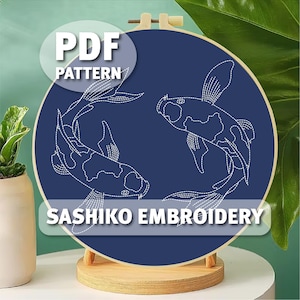 May include: A navy blue embroidery hoop featuring a Sashiko embroidery pattern of two koi fish. The design is stitched in white thread. The text "PDF PATTERN" and "SASHIKO EMBROIDERY" are displayed on the image.