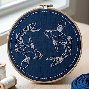 May include: Embroidered artwork featuring two koi fish in white thread on a navy blue fabric. The design is set within a wooden embroidery hoop. The fish are detailed with intricate stitching, creating a textured effect. The artwork is a decorative piece.