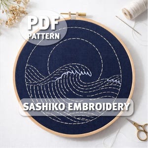 May include: A navy blue fabric stretched in a wooden embroidery hoop, featuring a white Sashiko embroidery design of a wave. The text "PDF PATTERN" and "SASHIKO EMBROIDERY" are overlaid on the image. A needle, thread, and scissors are also visible.