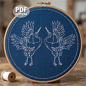 Peut inclure: Un cercle de broderie avec deux grues blanches brodées sur un tissu bleu marine. Les grues se font face, les ailes déployées. Le cercle est en bois, et le texte "PDF PATTERN" est visible en haut à gauche.