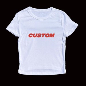 Op de afbeelding: Een wit t-shirt met het woord "CUSTOM" in rode letters op de borst. Het shirt heeft korte mouwen en een ronde hals. De achtergrond is effen zwart.