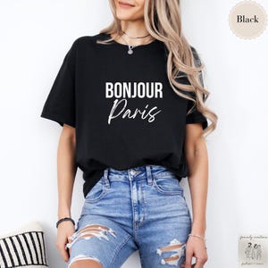 May include: Black t-shirt with the words "BONJOUR Paris" in white lettering. The shirt is a classic crew neck style. The model is wearing blue jeans.