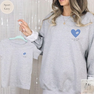 Peut inclure: Un sweat-shirt gris avec un cœur bleu contenant une pièce de puzzle et le texte "mama est. 2023". Un t-shirt gris avec une pièce de puzzle bleue et le texte "son est. 2023" est suspendu à un cintre.