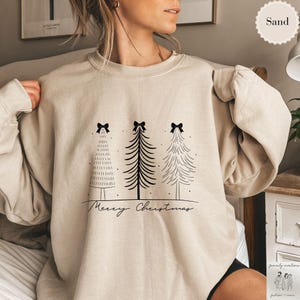 May include: Sand-colored sweatshirt featuring three stylized Christmas trees with black bows and a "Merry Christmas" script. The trees are in black and white, creating a minimalist holiday design. A cozy and festive apparel item.