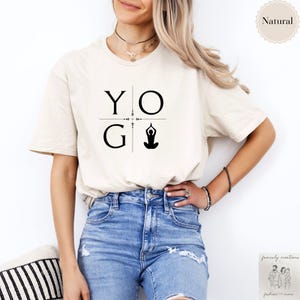May include: A cream-colored t-shirt with the word "YOG" in black block letters, with a yoga pose graphic. The shirt is short-sleeved and has a relaxed fit. The word "Natural" is printed on a circular label.