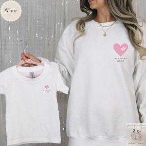 Pode incluir: Duas camisetas brancas com um coração rosa e uma peça de quebra-cabeça. A camiseta de adulto diz "mama est. 2023" e a camiseta de criança diz "livia est. 2023".