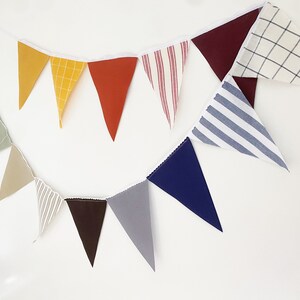 May include: A colorful fabric banner with triangular pennants in various colors and patterns, including yellow, orange, red, striped, and checkered designs. The banner is strung on a white cord, perfect for festive decorations.