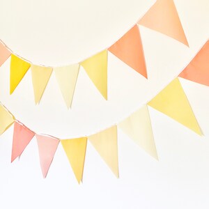 May include: A string of yellow, orange, and pink triangle flags strung together. The flags are made of paper and are hanging from a white wall.