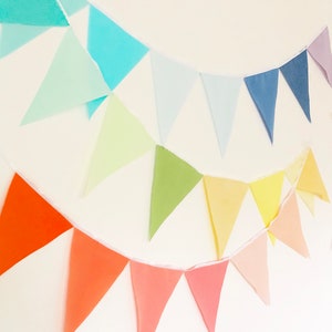 May include: A colorful fabric bunting with triangular flags in shades of blue, green, yellow, orange, and pink. The bunting is strung on a white cord and is hanging on a white wall.