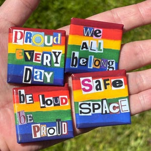 Puede incluir: Cuatro chapas cuadradas con un fondo de rayas arcoíris. Las chapas muestran texto en estilo de nota de rescate: PROUD EVERY DAY, WE ALL BELONG, BE LOUD BE PROUD y SAFE SPACE. Cada chapa tiene un acabado brillante y letras coloridas.