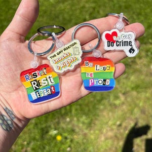 May include: Three acrylic keychains with metal rings. One features a rainbow background with the text "Resist, Rest, Repeat". Another is a white shape with "The Gay Agenda: Snacks & Rights" and a checklist. The third is a white shape with a red heart and "Be Gay Do Crime".