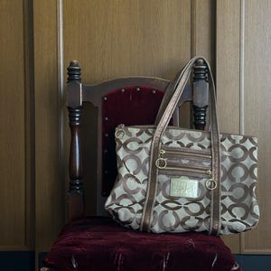 May include: A brown and tan tote bag with a repeating logo pattern, resting on a vintage wooden chair with a burgundy velvet seat. The bag has multiple zippered compartments and gold-toned hardware.