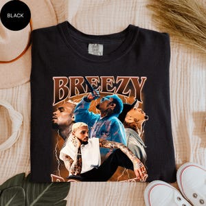 Puede incluir: Camiseta negra con un gr&aacute;fico de un cantante y la palabra "Breezy" en fuente marr&oacute;n. El gr&aacute;fico incluye im&aacute;genes del cantante en diferentes poses. La camiseta est&aacute; hecha de un material suave.