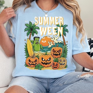 May include: Light blue t-shirt with a colorful graphic that says "SUMMER WEEN". The design features a beach scene with pumpkins, palm trees, a monster holding a surfboard, and ghosts. A fun, festive tee for Halloween.