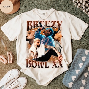 Puede incluir: Camiseta de color marfil con un gr&aacute;fico de un cantante, la palabra "Breezy" arriba y "Bowl XX" abajo. El gr&aacute;fico incluye im&aacute;genes del cantante en varias poses, con acentos de rayos.
